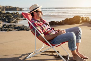 Yandan bakan olgun bir adam, yalnız oturuyor, bira tutuyor, kumsalın ve doğanın tadını çıkarıyor.