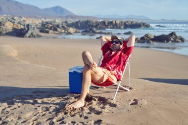 Olgun bir adam tek başına oturuyor, elleri başının üstünde. Kumsalın ve doğanın tadını çıkarıyor.
