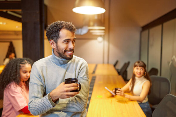 молодой латиноамериканец в офисе держит чашку кофе и улыбается