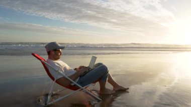 Genç adam tropikal bir plajda dizüstü bilgisayarla çalışıyor. Serbest ve iş konseptinde.