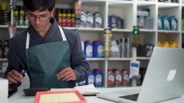 Genç erkek esnaf araba yedek parça dükkanının tezgahına hesap yapıyor ve notlar yazıyor.