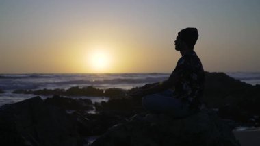 Genç adam gün batımında deniz kıyısındaki kayalarda meditasyon yapıyor.
