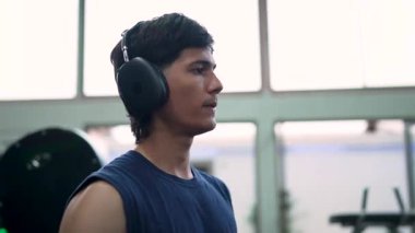 Kulaklıklı genç bir erkek sporcu spor aletlerini kullanarak egzersiz yaptıktan sonra kendini siliyor.