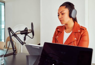 Genç bir kadın kulaklık takarak ses kaydı yapıyor ve stüdyoda mikrofona konuşuyor.