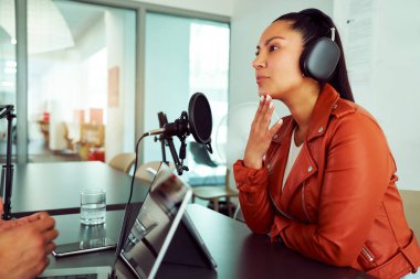 Kulaklık takan radyo sunucusu mikrofona konuşuyor ve prodüktörle modern stüdyoda podcast yapıyor.