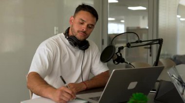 Stüdyoda dizüstü bilgisayar kullanarak ve notlar alarak odaklanan erkek podcaster