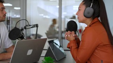 İki radyo sunucusu bir stüdyoda canlı yayın sırasında sohbet ediyorlar. Masada oturan kulaklık takan erkek ve kadın kayıt için dizüstü bilgisayar ve tablet bilgisayar kullanıyor. 