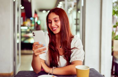 Kafede gülümseyen ve akıllı telefon kullanan genç bir kadın, cep telefonu teknolojisi ve çevrimiçi bağlantının keyfini çıkarıyor