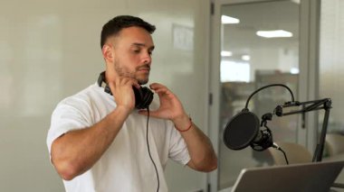 Beyaz tişörtlü bir adam stüdyoda dizüstü bilgisayar kullanarak ses yayını yapıyor ve konuğunu dinliyor. 