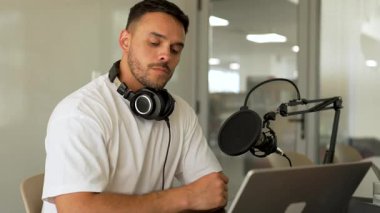 Beyaz tişörtlü bir adam stüdyoda dizüstü bilgisayar kullanarak ses yayını yapıyor ve konuğunu dinliyor. 