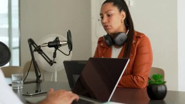 İki radyo sunucusu bir stüdyoda canlı yayın sırasında sohbet ediyorlar. Masada oturan kulaklık takan erkek ve kadın kayıt için dizüstü bilgisayar ve tablet bilgisayar kullanıyor. 