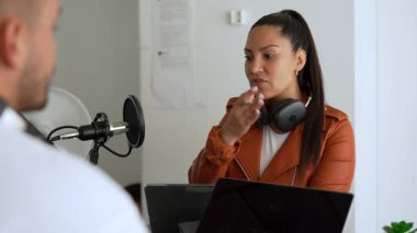 İki radyo sunucusu bir stüdyoda canlı yayın sırasında sohbet ediyorlar. Masada oturan kulaklık takan erkek ve kadın kayıt için dizüstü bilgisayar ve tablet bilgisayar kullanıyor. 