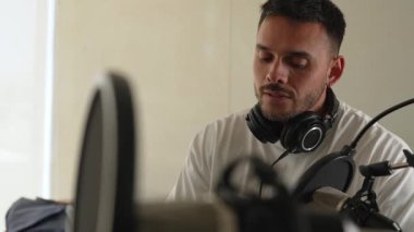 Beyaz tişörtlü bir adam profesyonel ekipman kullanarak stüdyoda ses yayını yapıyor.