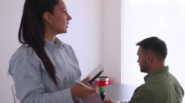 İki iş arkadaşı bir projeyi tartışıyor, fikirleri paylaşıyor, kadın tablet bilgisayarla ayakta duruyor, adam dizüstü bilgisayarla masada oturuyor.