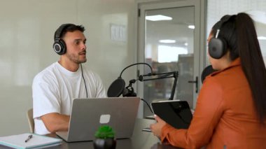 İki radyo sunucusu bir stüdyoda canlı yayın sırasında sohbet ediyorlar. Masada oturan kulaklık takan erkek ve kadın kayıt için dizüstü bilgisayar ve tablet bilgisayar kullanıyor. 