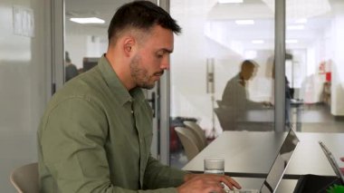 Genç adam ofiste dizüstü bilgisayar kullanarak masada oturuyor.