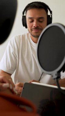 Beyaz tişörtlü bir adam stüdyoda dizüstü bilgisayar kullanarak ses yayını yapıyor ve konuğunu dinliyor. 