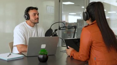 İki radyo sunucusu bir stüdyoda canlı yayın sırasında sohbet ediyorlar. Masada oturan kulaklık takan erkek ve kadın kayıt için dizüstü bilgisayar ve tablet bilgisayar kullanıyor. 