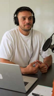 Beyaz tişörtlü bir adam dizüstü bilgisayar kullanarak stüdyoda ses yayını yapıyor.