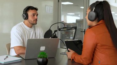 İki radyo sunucusu bir stüdyoda canlı yayın sırasında sohbet ediyorlar. Masada oturan kulaklık takan erkek ve kadın kayıt için dizüstü bilgisayar ve tablet bilgisayar kullanıyor. 