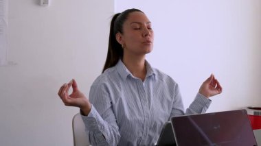 Genç iş kadını ofisinde meditasyon yapıyor. Masasında tabletinin önünde oturuyor. 