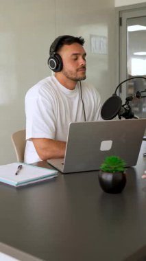 Beyaz tişörtlü bir adam stüdyoda dizüstü bilgisayar kullanarak ses yayını yapıyor ve konuğunu dinliyor. 