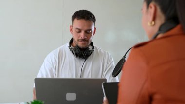 İki radyo sunucusu bir stüdyoda canlı yayın sırasında sohbet ediyorlar. Masada oturan kulaklık takan erkek ve kadın kayıt için dizüstü bilgisayar ve tablet bilgisayar kullanıyor. 