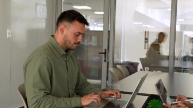 Genç bir adam masanın başında dizüstü bilgisayarla oturuyor. Ofisteki bayan meslektaşının önünde.