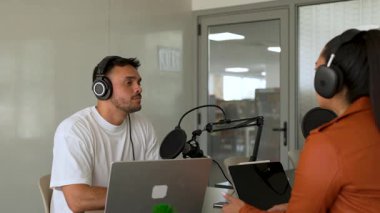 İki radyo sunucusu bir stüdyoda canlı yayın sırasında sohbet ediyorlar. Masada oturan kulaklık takan erkek ve kadın kayıt için dizüstü bilgisayar ve tablet bilgisayar kullanıyor. 