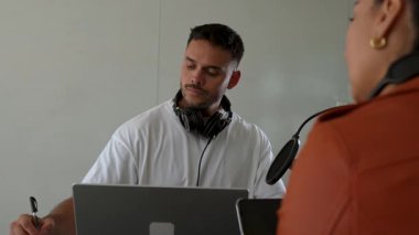 İki radyo sunucusu bir stüdyoda canlı yayın sırasında sohbet ediyorlar. Masada oturan kulaklık takan erkek ve kadın kayıt için dizüstü bilgisayar ve tablet bilgisayar kullanıyor. 