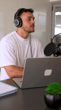 Beyaz tişörtlü bir adam stüdyoda dizüstü bilgisayar kullanarak ses yayını yapıyor ve konuğunu dinliyor. 