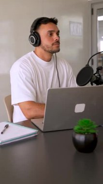 Beyaz tişörtlü bir adam stüdyoda dizüstü bilgisayar kullanarak ses yayını yapıyor ve konuğunu dinliyor. 