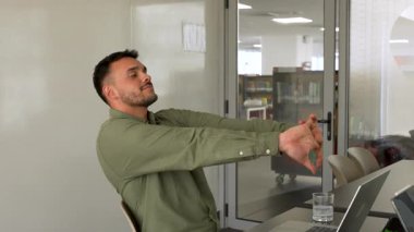 Genç adam masada oturuyor ve ofiste dizüstü bilgisayar kullanmadan önce geriniyor.