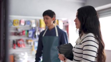 İki genç çalışan, bir erkek ve bir kadın, bir tablet kullanarak bir nalbur dükkanında envanter yönetimi yapıyorlar.