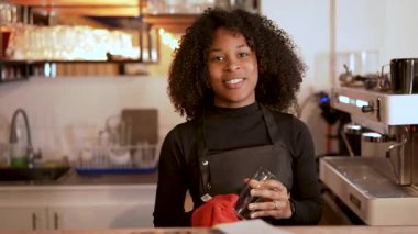 Genç gülümseyen Afrikalı Amerikalı barmen bir kafede kırmızı bir bezle bardağı parlatıyor.