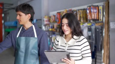 İki genç çalışan, bir erkek ve bir kadın, bir tablet kullanarak bir nalbur dükkanında envanter yönetimi yapıyorlar.