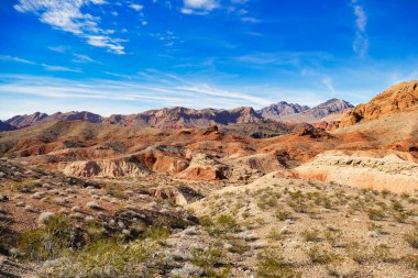 Kuru çöl manzarası aşınmış kırmızı kayalar, seyrek bitki örtüsü ve arka planda karanlık dağlar, Ateş Vadisi Parkı, Nevada, ABD.