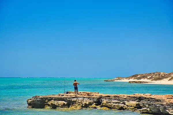 stock image Dark skinned fisherman on a rock in the azure blue Indian Ocean, at the remote coast of Cape Range National Park, Western Australia