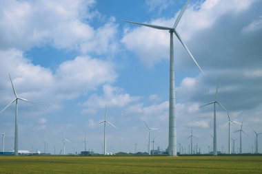 Düzinelerce büyük rüzgar türbini düz bir çayırda, arka planda endüstriyel binalar. Eemshaven, Groningen ili, Hollanda.
