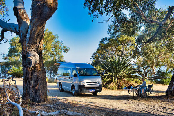 Small motorhome and elderly woman sitting in a camping chair at a beautiful camping spot on the shore of Lake Indoon, shire of Carnamah, west coast of Western Australia.