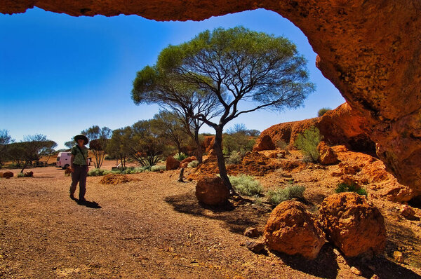 Woman walking to a natural arch of red sandstone in the desert of the Australian outback. London Bridge, shire of Sandstone, Western Australia