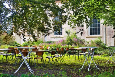 Empty wooden office chairs and tables in a garden for an informal social gathering in the shade of an old beech tree. No people. Wall of an old church in the background. Warffum, province of Groningen, Netherlands clipart