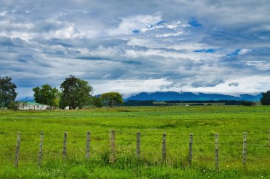 Yeşil çayırlar, eski çitler, basit çiftlik evleri ve dramatik bir gökyüzünün altındaki karanlık tepeler. Manawatu bölgesi, Kuzey Adası, Yeni Zelanda, Fielding 'e yakın..