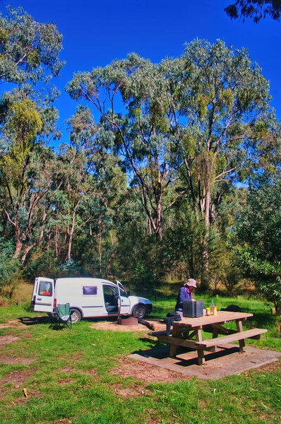 Middle aged woman with a small campervan preparing food at a camping table in an eucalyptus forest. Burrowa-Pine National Park, Victoria, Australia