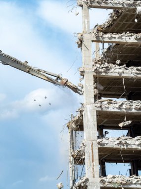 Ağır makineler çok katlı bir binayı etkin bir şekilde yıkıyor ve yapıdan enkaz dökülüyor. Parlak gökyüzü, devam eden inşaat çalışmalarını gözler önüne seren tozlu manzaraya zıtlık sağlıyor..