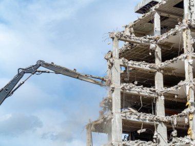 Büyük bir yıkım makinesi bulutlu bir gökyüzü altında yüksek bir binayı yıkıyor. Kentsel bir alanda bina çökmeye başladığında toz ve enkaz görülebilir..