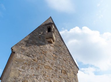 Eski taş yapıda sivri bir çatı ve küçük bir pencere var. Açık mavi bir gökyüzüne karşı duruyor, sakin bir manzara sergiliyor. Sahne bir sükunet ve tarih anı yakalıyor..