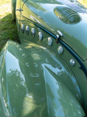 Çimenlerin üzerine park edilmiş eski yeşil bir araba zarif kıvrımlarını ve parlak krom aksanlarını sergiler. Güneş ışığı parlak yüzeyinden yansıyarak klasik görünüşünü geliştirir..