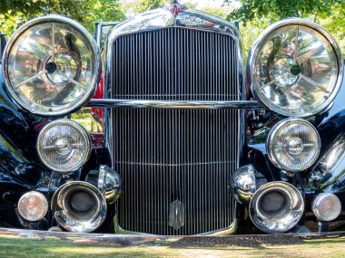 Eski model bir araba, parlak krom özelliklerini ve büyük farlarını bir araba fuarında gururla sergiler. Aydınlık bir günde yemyeşil ortamda katılımcılar araca hayran..