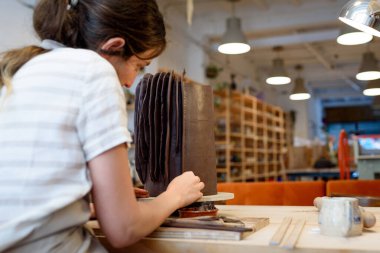 Çömlek atölyesinde çiğ seramik vazo ve kilden esrar yapan bir kadın. Doğal ışık kazıma, düzleştirme, şekillendirme ve yontma ile çömlek atölyesi. Kil tenceresi yapmak